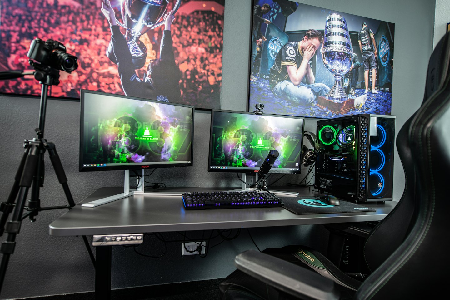 Dual monitors on a dark desk with gaming PC and camera.